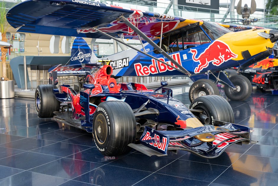 A detailed image of a Red Bull racing car and a Red Bull-branded aircraft displayed indoors, likely within a modern exhibition or showroom with a tiled black floor and glass walls. The racing car, positioned in the foreground, is a single-seater with a sleek, aerodynamic body, large black racing tires, and sponsor logos including Red Bull and Bridgestone. The aircraft, situated behind the car, is a biplane with a blue fuselage featuring prominent Red Bull branding, a yellow nose, and a large red bull logo on the side. The aircraft's propeller and supporting struts are visible, with part of the cockpit also in view. The background includes a reception area or bar counter with bottles and utensils, indicating an enclosed space designed for display or promotional events. The image captures the process of moving or transporting high-value objects like racing vehicles and aircraft, demonstrating professionally coordinated logistics suitable for a house or special event relocation. Man with Van Bulls Cross offers services that include the safe handling and transport of such large, delicate items.