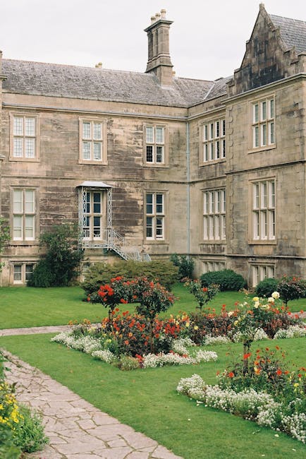 A historic stone-built mansion with multiple large sash windows and a steeply pitched slate roof, situated behind a well-maintained garden with colorful flower beds and neatly trimmed lawns. A metal staircase leads to a side door on the first floor, while a pathway runs alongside the garden. The scene appears to be outdoors during daytime with overcast skies. This image illustrates a typical setting for house removals or furniture transport, as a team from Man with Van Bulls Cross may be involved in packing, loading, or moving household items from such a property as part of a home relocation process. The stone façade and garden setting highlight the importance of careful planning and logistics in house removals services, particularly for period properties requiring specialist handling during packing and loading procedures.