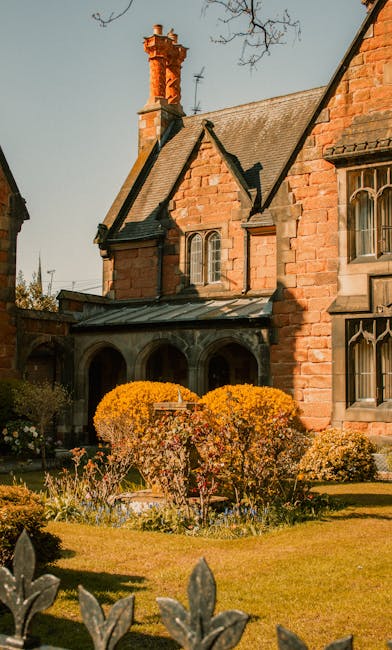 A historic stone-built mansion with multiple large sash windows and a steeply pitched slate roof, situated behind a well-maintained garden with colorful flower beds and neatly trimmed lawns. A metal staircase leads to a side door on the first floor, while a pathway runs alongside the garden. The scene appears to be outdoors during daytime with overcast skies. This image illustrates a typical setting for house removals or furniture transport, as a team from Man with Van Bulls Cross may be involved in packing, loading, or moving household items from such a property as part of a home relocation process. The stone façade and garden setting highlight the importance of careful planning and logistics in house removals services, particularly for period properties requiring specialist handling during packing and loading procedures.