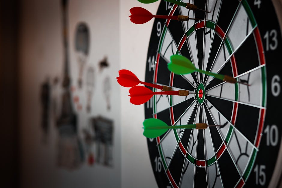 Inside a room with a standard dartboard mounted on the wall, the dartboard features black, white, green, and red sections with numbers around the edge. Several darts with red and green plastic fins are embedded in different areas of the dartboard, including the bullseye and outer scoring sections. The focus is on the dartboard, with some blurred objects visible on the beige wall behind it, such as hooks or small shelves. The environment appears well-lit with natural or ambient lighting. This setting could be part of a casual recreational area in a home, which might be considered during a home relocation or packing and moving process. The image naturally connects to the theme of pre-move activity or creating a functional space in a residence, relevant to the services offered by Man with Van Bulls Cross for house removals and moving logistics.