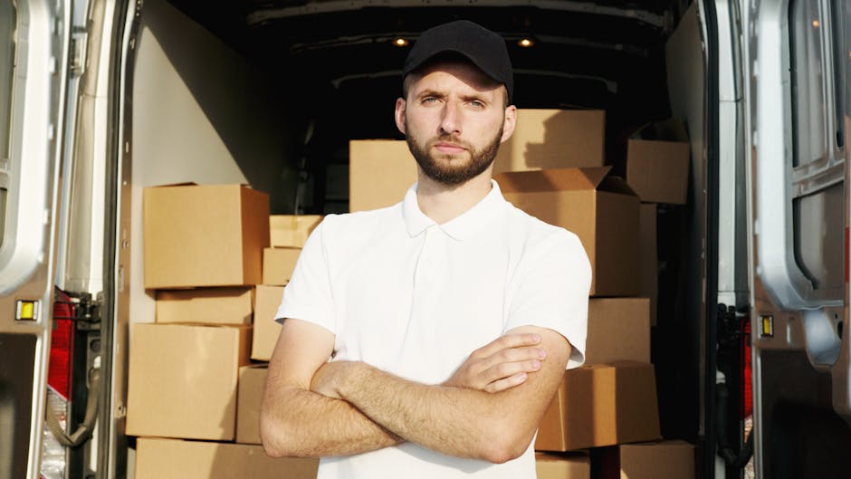 A young man with a short beard, wearing a white polo shirt and a black cap, stands with crossed arms inside the back of a moving van, which is loaded with numerous sealed cardboard boxes of various si
