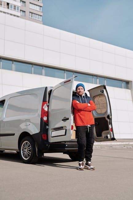 A young man with a short beard, wearing a white polo shirt and a black cap, stands with crossed arms inside the back of a moving van, which is loaded with numerous sealed cardboard boxes of various si