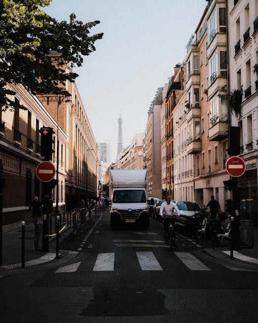 A white moving van is positioned in the middle of a narrow urban street during daytime, with its front facing the viewer. The vehicle is surrounded by multi-storey buildings with classic Parisian architecture, including balconies and large windows, lining both sides of the street. The road is marked with zebra crossings, and there are two red and white ‘no entry’ signs on either side of the van. The street appears to be temporarily closed to other traffic to facilitate home relocations or furniture transport, with a cyclist riding past the van and several pedestrians nearby. In the background, the Eiffel Tower is visible under a clear sky. The image captures the process of loading or unloading during a residential move, supported by [COMPANY_NAME] and their specialist moving services. The scene emphasizes city logistics related to house removals, with a focus on access and vehicle positioning in a narrow street on Whitewebbs, Bulls Cross.