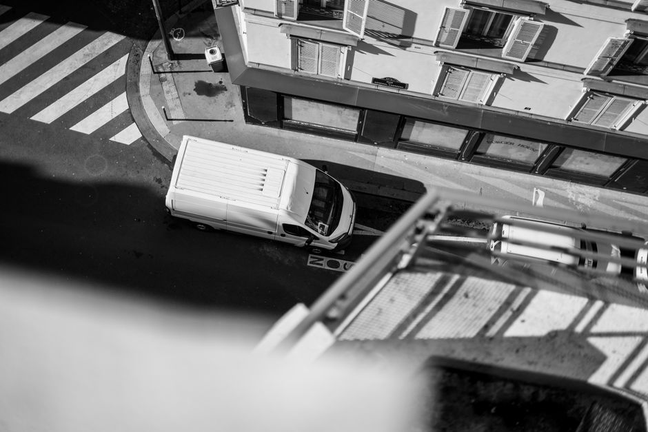 An overhead view of a narrow street in Whitewebbs, Bulls Cross, showing a white cargo van parked parallel to the sidewalk outside a residential building with multiple folding windows. The van is positioned near a pedestrian crosswalk with bold white and black striped markings, and a traffic sign is visible at the corner. To the right of the van, a portion of a steel balcony railing extends into the frame, casting shadows on the road. The building's exterior features several closed window shutters and a small air conditioning unit. This scene captures the logistics involved in house removals, with the van ready for loading or unloading household furniture and boxes, supported by Man with Van Bulls Cross' professional moving services, emphasizing careful furniture transport and packing during a home relocation on a narrow urban street.