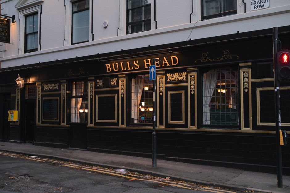 Photograph of the exterior of Bulls Head, a pub with dark green and gold ornate exterior decoration, situated on a street corner. The building has large windows with white curtains, through which warm indoor lighting is visible. A black sign with gold lettering displaying 'BULLS HEAD' is mounted above the windows and entrance. A blue one-way traffic sign is positioned on a pole in front of the pub. To the right, there is a pedestrian crossing signal showing a red figure. The sidewalk in front is clean with a few scattered leaves. The scene captures a moment in daytime with natural light, relevant to property or business relocation efforts such as packing or furniture transport associated with house removals near Bulls Cross, and reflects the kind of environment where [COMPANY_NAME] might assist with moving logistics.
