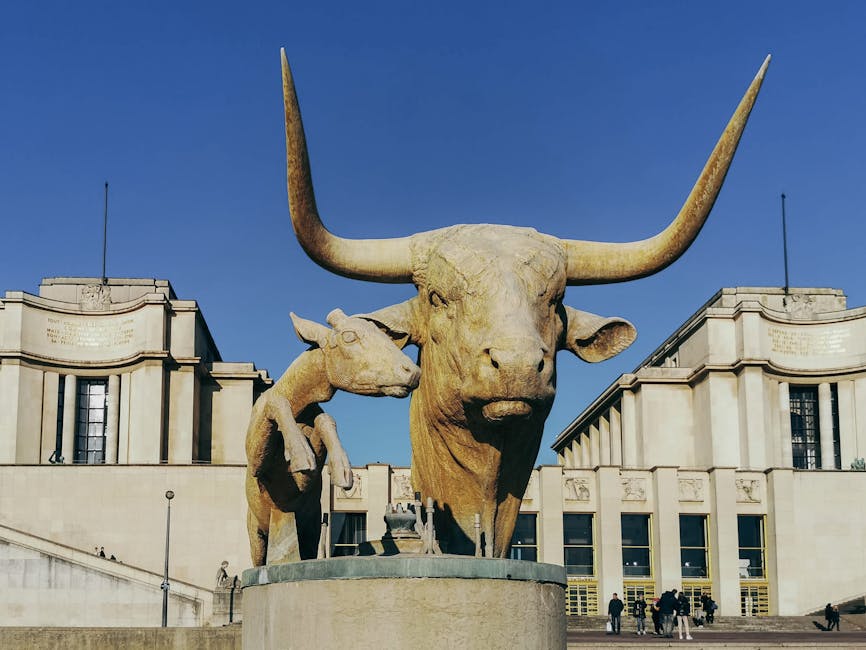 Photograph of the Wellington Arch in London with a large, prominent sculpture of two animals on a circular pedestal in the foreground. The sculpture features a large bull with long, upward-curving horns standing next to a smaller animal, possibly a calf or young bull. The scene is set outdoors on a bright, clear day with a blue sky. In the background, the architecture of the Wellington Arch building is visible, showcasing symmetrical, classical-style stone facades with decorative panels and tall windows. The area around the sculpture and building appears to be a public space with a few people walking or standing nearby. This setting exemplifies an urban environment often associated with city landmarks, and the sculpture is made of a stone material with detailed features. The context suggests an emphasis on area aesthetics, possibly related to city planning or public art installed within the vicinity of notable landmarks, relevant to the topic of furniture transport and home relocation services offered by Man with Van Bulls Cross.