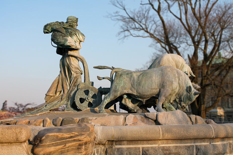 Photograph of the Wellington Arch in London with a large, prominent sculpture of two animals on a circular pedestal in the foreground. The sculpture features a large bull with long, upward-curving horns standing next to a smaller animal, possibly a calf or young bull. The scene is set outdoors on a bright, clear day with a blue sky. In the background, the architecture of the Wellington Arch building is visible, showcasing symmetrical, classical-style stone facades with decorative panels and tall windows. The area around the sculpture and building appears to be a public space with a few people walking or standing nearby. This setting exemplifies an urban environment often associated with city landmarks, and the sculpture is made of a stone material with detailed features. The context suggests an emphasis on area aesthetics, possibly related to city planning or public art installed within the vicinity of notable landmarks, relevant to the topic of furniture transport and home relocation services offered by Man with Van Bulls Cross.