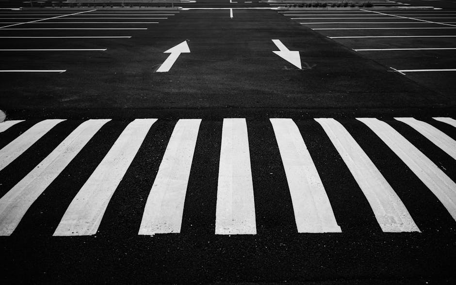 A black and white image of a parking lot entrance featuring a pedestrian crossing with white striped lines in the foreground. Beyond the crossing, there are two lanes with painted white arrows indicating the direction of traffic flow, one pointing straight ahead and the other pointing left. The surface of the lot appears smooth and well-maintained, with no vehicles visible. This scene is associated with logistical planning for home relocations or furniture transport, as part of the services offered by Man with Van Bulls Cross, which involves careful management of parking access and loading areas for efficient moving and packing processes.