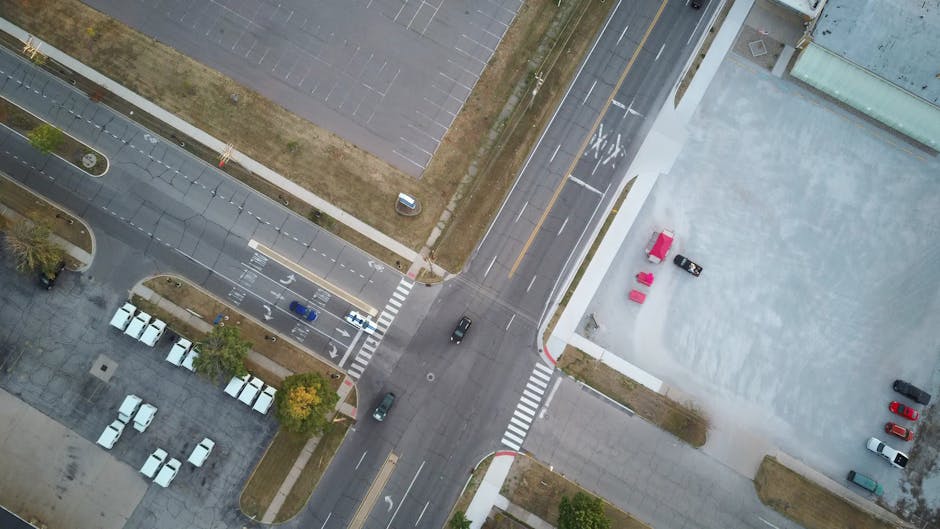 An aerial view demonstrating the intersection of Whitewebbs Lane with parking areas and adjacent buildings in Bulls Cross. In the top left corner, a section of parking lots with marked spaces and several white vehicles are visible, along with indicating paved walkways and some greenery. To the right, there is a large, grey, open concrete area with a few parked cars and pink outdoor furniture positioned near a white building. Below, a section of road with vehicle lanes, crosswalks, and some moving cars can be seen, along with signs and parking restrictions. The image captures the outdoor environment and logistics involved in house removals, illustrating the importance of accessible parking and clear routes for smooth furniture transport and home relocation processes, as managed by Man with Van Bulls Cross.